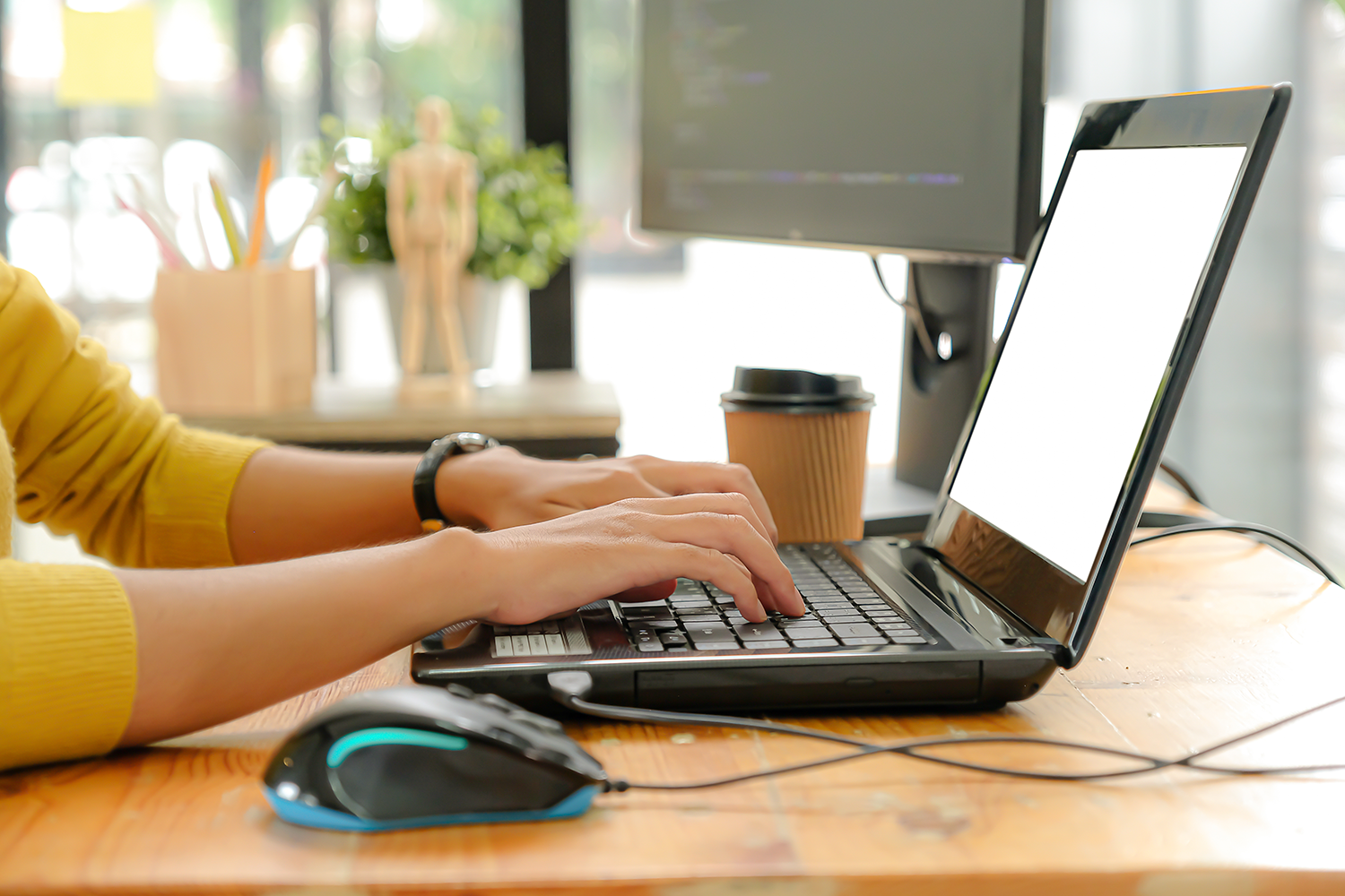 Personne tapant sur un ordinateur portable à un bureau en bois avec une souris, une tasse de café et un moniteur dans un espace de travail moderne et lumineux, idéal pour préparer des présentations Wordcamp.